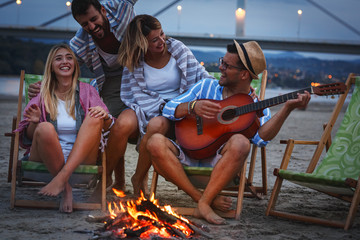 Group of friends sitting around camp fire at the beach at the autumn evening.They play guitar and...