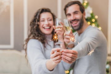 Loving and stylish couple in holiday atmosphere, standing with bengal lights, smiling and cheerring. Focus on the sparkles on the forefront.