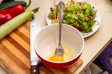 Smashing chicken egg into a red bowl in the kitchen with a knife and some vegetables on a table