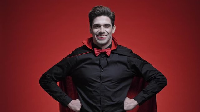 Vampire man with blood and fangs with hands on hips in black halloween costume is smiling isolated over red wall