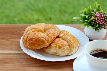Croissant on white plate and cup of hot drink coffee on wooden table.