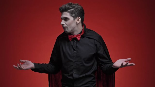 Walking vampire man with blood and fangs in black halloween costume looking for something and feeling disappointed isolated over red wall