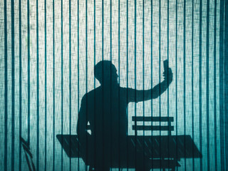 silhouette on turquoise office curtain of a sitting man taking a self-portrait with his smartphone