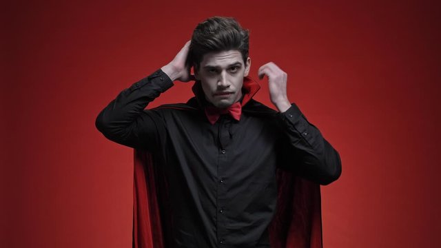Vampire man with blood and fangs in black halloween costume is fixing his bow tie, collar, hair and teeth isolated over red wall