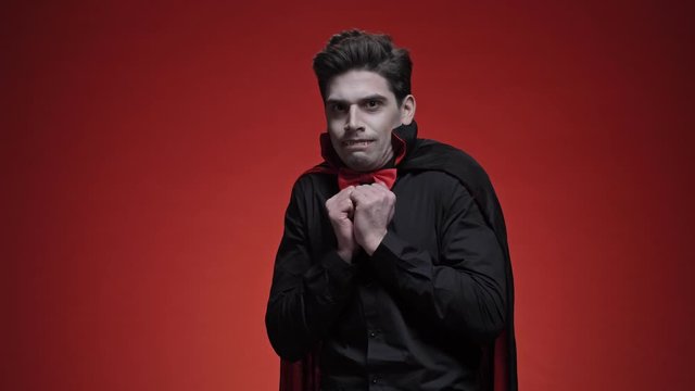 Vampire man with fangs in black halloween costume becoming scared isolated over red wall looking at the camera