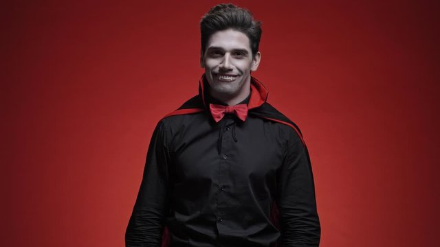 Happy and smiling vampire man with blood and fangs in black halloween costume waving hand to the camera isolated over red wall