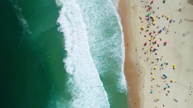 Aerial locked shot of waves breaking on the shore. Colourful beach umbrellas and people enjoying the summer. 4K.