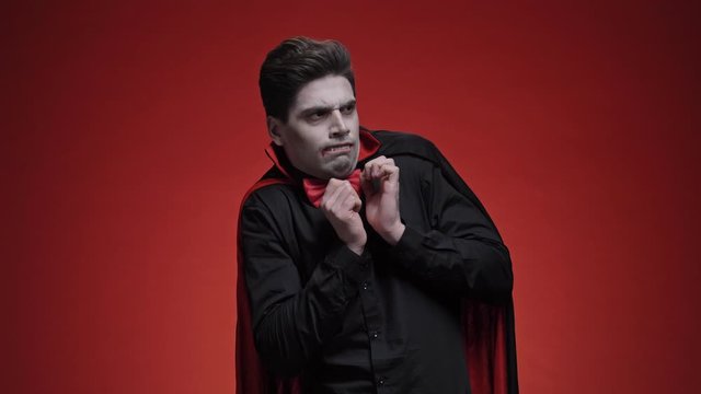 Displeased vampire man with fangs in black halloween costume becoming scared of something while looking away isolated over red wall