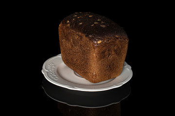 One whole fresh baked dark bread on white plate isolated on black glass