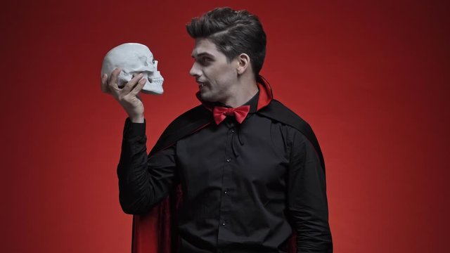 Scary vampire man with fangs in black halloween costume holding human skull and turning it to the camera isolated over red wall