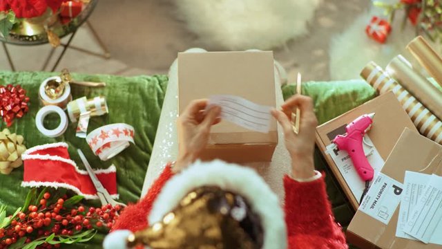 Holiday season. Upper view of stylish woman in sparkle gold sequin santa hat sitting on sofa in the modern living room at Christmas sending parcels. This video was shot in PRORes 422 codec.