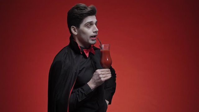 Glad vampire man with fangs in black halloween costume holding a tomato juice and drinking it with a cocktail tube isolated over red wall