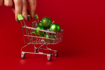 green Christmas toys lie in a small supermarket trolley on a red background. Female hands hold the trolley. Christmas shopping concept on a red background.