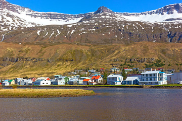 Landscape of coastal town in Northern Iceland during spring. Mountain tops covered in snow, while waterfalls located beneath them rushing towards the sea waters.