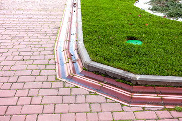 Curved drainage system in the park by the green lawn and footpath of red paving slabs.