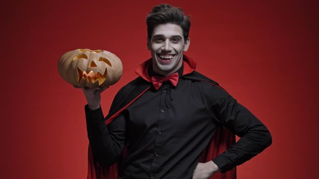 Vampire man with blood and fangs in black halloween costume congratulates with pumpkin in his hands isolated over red wall