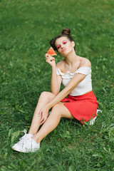 Fototapeta premium A young girl with a cheerful hairstyle and bright makeup holds a piece of watermelon in her hands and sits on the green grass, lips folded in the form of a kiss