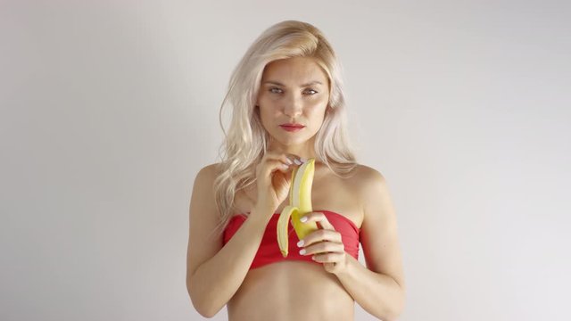 Waist-up shot of young Caucasian woman with long blonde hair, in red strapless bikini top, standing against white wall in studio and peeling banana while looking straight at camera