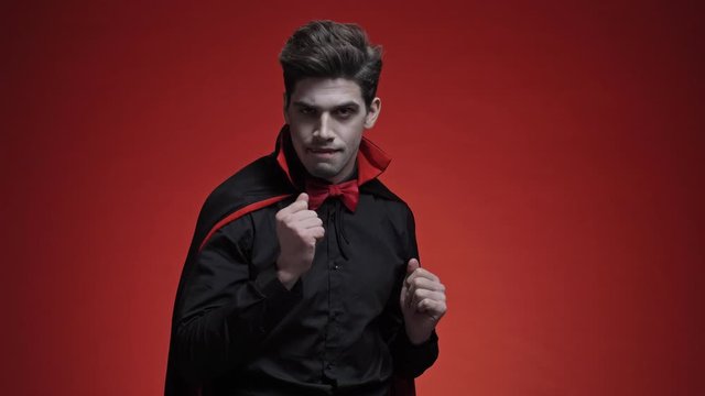 Vampire man with blood and fangs in black halloween costume is dancing and smiling isolated over red wall