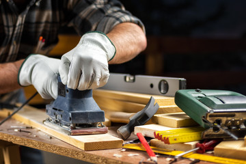 Adult carpenter craftsman wearing leather protective gloves, with the electric sander smoothes a wooden table. Construction industry, housework do it yourself. Safety at work.