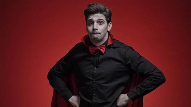 Vampire man with fangs in black halloween costume becoming confused and shrugging his shoulders isolated over red wall