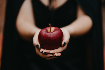 Black witch holding red apple in hands on autumn forest background. Horror, halloween, cosplay...
