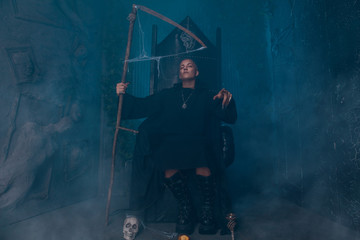 young very angry man with a caucasian face is dressed in a black robe and leather big boots and sits with scythe on scary vampire throne in the dark at Halloween night.