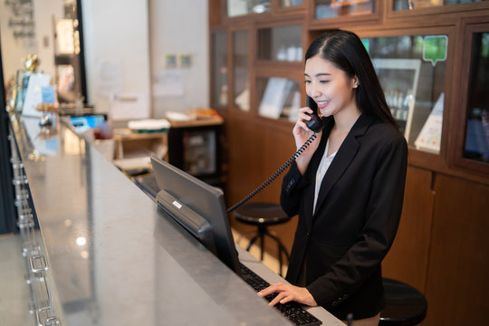 Welcome to the hotel,Happy young Asian woman hotel receptionist worker smiling standing,she taking  telephone call at a Modern luxury reception counter waiting for guests getting key card in hotel