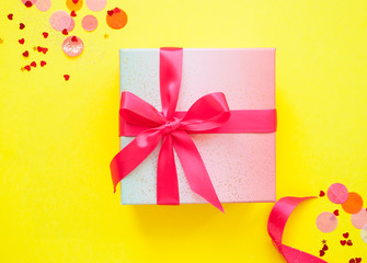 Giftbox tied with pink color ribbon on bright yellow background with sparklets and confetti. Flat lay style.