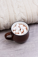 Fototapeta na wymiar Cozy winter home background, cup of hot cocoa with marshmallow, warm knitted sweater on white wooden board background.