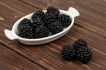 Lot of whole fresh black blackberry in white oval ceramic bowl on brown wood