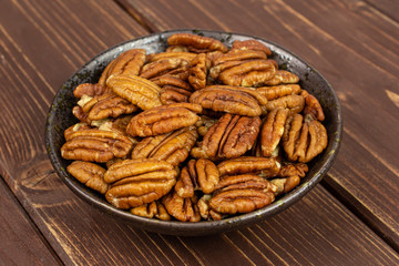 Lot of whole dry brown pecan nut in dark ceramic bowl on brown wood