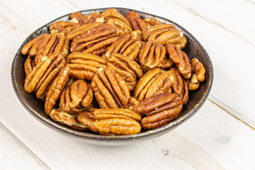 Lot of whole dry brown pecan nut in dark ceramic bowl on white wood