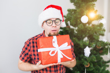 Christmas and holidays concept - Funny man in santa hat holding a gift at home in the living room