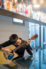 Young Asian man guitarist playing acoustic guitar at bar counter in the cafe. Practicing string musical instrument. Music and entertainment concept.