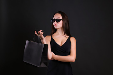 Fototapeta na wymiar Black friday sale concept. Attractive young woman with long brunette hair, smiling, wearing sexy dress & cat eye sunglasses, holding blank shopping bag over black background. Copy space, close up.