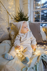 Christmas morning. A child in a white suit sits joyful on the bed. Around him are light bulbs, garlands