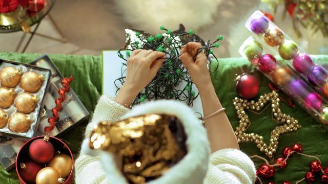 Holiday season. Upper view of elegant woman in sparkle gold sequin santa hat sitting on sofa in the modern living room at Christmas trying to untangle tangled christmas lights. PRORes 422 codec.