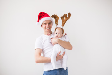 Fototapeta premium My first Christmas. Dad in santa hat holding his baby girl daughter wearing in deer horns on white background with copy space