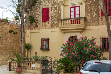 Fototapeta na wymiar Picturesque house in Mdina, Malta, with brightly red door and windows' frames and shutters, and with lush Nerium oleander bush growing near the fence door gate.