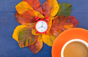 Change The Clock Winter Time Concept or Autumn Morning Mood. Creative Top View Flat Lay Composition With Red Alarm Clock, Bright Fall Leaves and Orange Cup of Coffee Latte are on Ink Blue Wooden Desk.