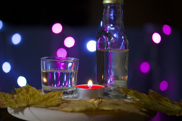 Obraz na płótnie Canvas Burning candle with a drink on the background with colorful lights. Nearby is a glass and a bottle of drink. Laid out autumn maple leaves. Holiday accessories at dusk.