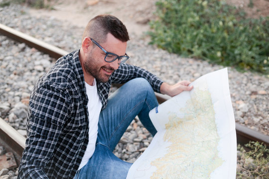 Chico joven sentado en las v&iacute;as del tren ojeando un mapa de viajes al atardecer