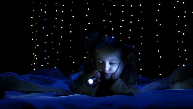 Little girl on the bed and shines a flashlight at the book she reads. Bokeh background. Slow motion