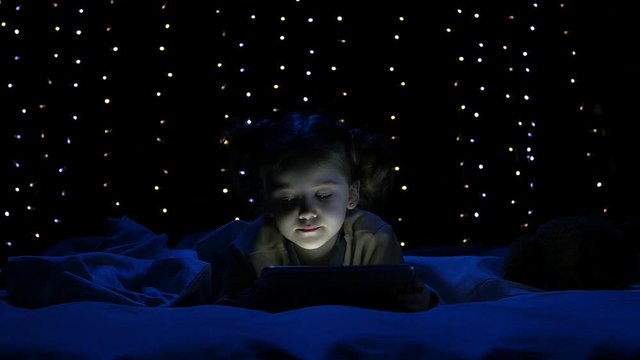 Child is lying on a bed holding a laptop and watching a cartoon. Bokeh background. Slow motion