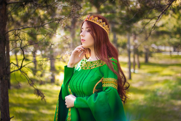 A beautiful, young, red-haired girl in a green medieval dress with long sleeves, with a golden crown, standing on the grass, in a fairy forest. Fantastic photosession. A model with clean skin.