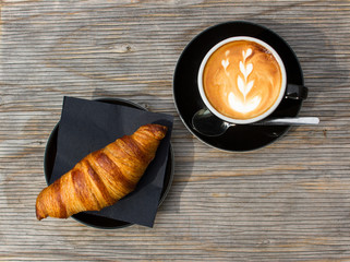 Flatlay of a cappuccino with beautiful latte art and a delicious croissant on a wooden table