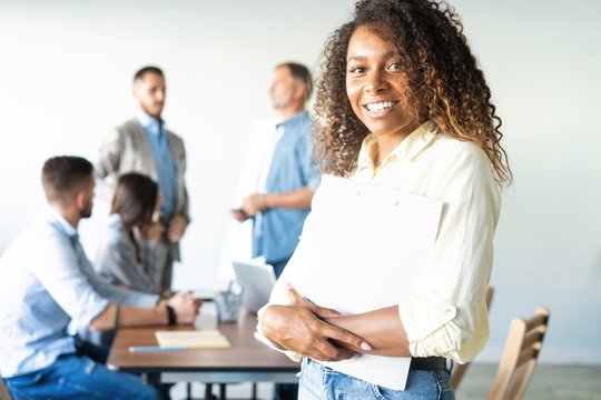 Beautiful young professional african woman standing in office while workers hold a meeting in background.
