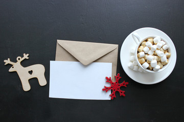 A Cup of cocoa with marshmallows and an empty envelope on the background of a Christmas tree and Christmas tree toys, a wooden deer, cinnamon sticks. Christmas background, bokeh, copy space.