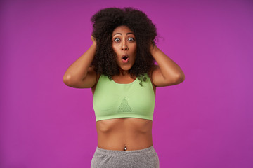 Obraz premium Amazed young curly dark skinned female wearing light green top, holding hea with hands and looking at camera with round eyes and opened mouth, isolated over purple background